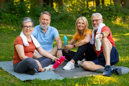 Téléchargez les photos : Des amis aînés souriants avec des bouteilles d'eau reposant sur des tapis de yoga après une formation en plein air, des hommes et des femmes âgés heureux se détendre dans un parc verdoyant, profiter de l'autre compagnie, représentant un mode de vie sain - en image libre de droit