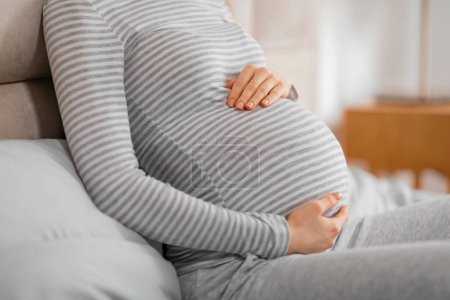 Téléchargez les photos : Femme enceinte dans un cadre confortable à la maison, reposant doucement ses mains sur sa chemise rayée recouverte de bosse de bébé - en image libre de droit