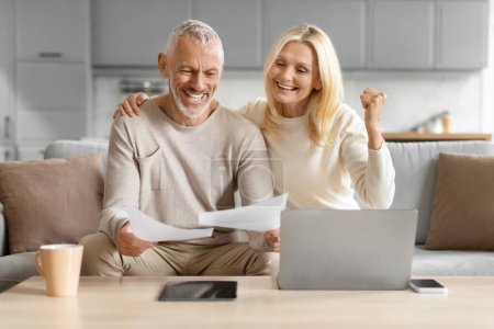 Alegre pareja de ancianos celebrando las buenas noticias con el papeleo en la mano, sentado junto a un ordenador portátil en casa