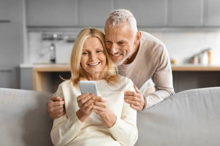 Téléchargez les photos : Couple âgé joyeux s'engageant avec un smartphone, montrant un moment de connectivité et de plaisir à la maison - en image libre de droit