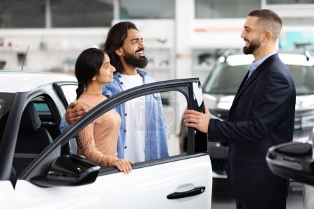 Foto de Un vendedor sonriente abre la puerta de un nuevo vehículo para los clientes pareja india en una sala de exposición de coches, que ofrece una vista interior - Imagen libre de derechos
