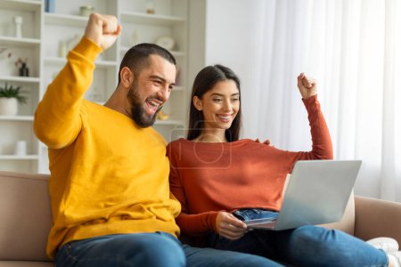 Photo for A joyful man and woman celebrating successful event or victory, looking at a laptop screen, raising their fists in excitement - Royalty Free Image