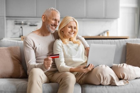 Photo for Smiling mature couple using a laptop and credit card, appearing to be making an online purchase while seated on a sofa - Royalty Free Image