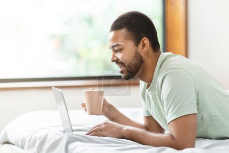 Photo for A relaxed setting with african american man in casual attire lying on his bed, absorbed in his laptop screen while holding a warm beverage - Royalty Free Image