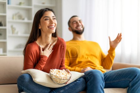 Téléchargez les photos : Un homme et une femme joyeux assis sur un canapé, la femme tenant un bol de pop-corn, tous deux semblent s'amuser à regarder la télévision - en image libre de droit
