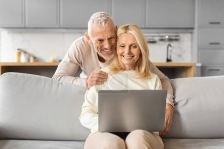 Téléchargez les photos : Couple âgé affectueux assis sur un canapé tout en regardant un écran d'ordinateur portable, partageant un moment - en image libre de droit