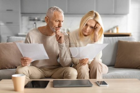 Reifes Paar schaut besorgt auf Dokumente, sitzt mit Tablet und Kaffeetasse auf dem Sofa