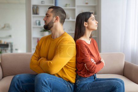 Ein junger Mann und eine junge Frau sitzen Rücken an Rücken auf einer Couch und schauen beide mit aufgeregten Gesichtsausdrücken weg, was eine Meinungsverschiedenheit impliziert.
