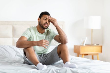 Foto de Hombre afroamericano se sienta en la cama sosteniendo un vaso de agua, con aspecto cansado o enfermo en un ambiente cómodo en casa - Imagen libre de derechos