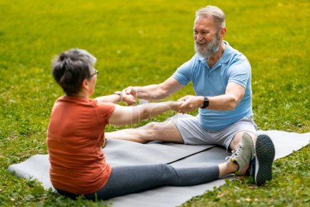 Téléchargez les photos : Couple âgé souriant s'engageant dans des exercices d'étirement partenaire sur tapis de yoga à l'extérieur, homme et femme âgés faisant de l'exercice ensemble dans un parc vert animé, se tenant la main, mettant en valeur la coopération et la forme physique - en image libre de droit