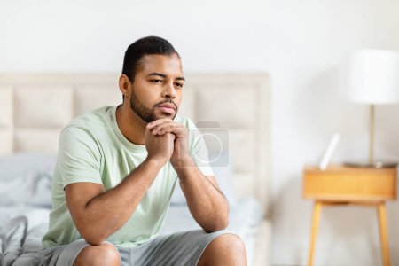 Téléchargez les photos : Un homme noir réfléchi est capturé assis sur un lit regardant loin avec une main sur son menton, dans un cadre de chambre - en image libre de droit