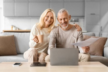 Älteres Paar liest aufgeregt ein Dokument mit Kaffee in der Hand und sitzt zu Hause neben einem Laptop