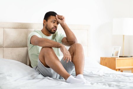 Foto de Un hombre afroamericano pensativo sentado en su cama, apareciendo perdido en el pensamiento, con un fondo de enfoque suave - Imagen libre de derechos