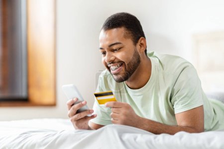 Téléchargez les photos : Jeune homme afro-américain tenant une carte de crédit et regardant son smartphone, effectuant probablement un achat en ligne - en image libre de droit