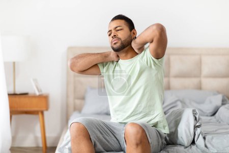 Foto de Hombre afroamericano hace muecas con malestar mientras toca su cuello, sentado en la cama en un ambiente de dormitorio minimalista - Imagen libre de derechos