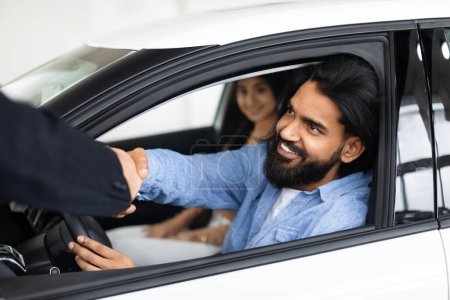 Téléchargez les photos : Un client indien joyeux dans une voiture serrant la main d'un vendeur de voitures à travers une fenêtre ouverte dans un concessionnaire - en image libre de droit