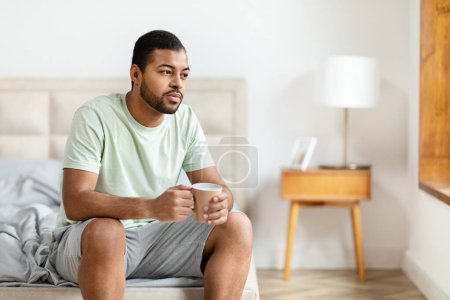 Foto de Hombre afroamericano se sienta en el borde de una cama sosteniendo una taza de café, en un dormitorio bien iluminado con una expresión serena - Imagen libre de derechos