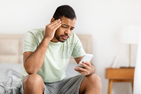 Photo for African american man appears stressed and concerned while looking at a smartphone, sitting on a bed in a minimalistic bedroom - Royalty Free Image
