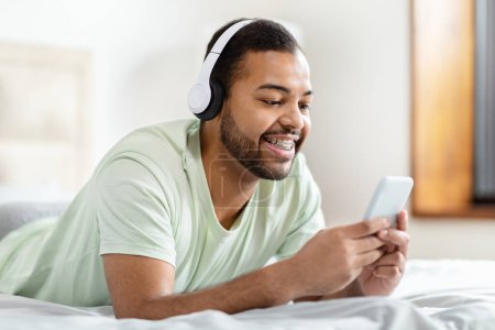 Photo for Casual african american man in bed with headphones browsing on his smartphone with focus and enjoyment - Royalty Free Image