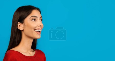 Foto de Retrato de una joven exuberante en suéter rojo, mirando a un lado con una amplia sonrisa, mujer milenaria emocionada de pie aislada sobre un fondo de estudio azul claro, panorama - Imagen libre de derechos