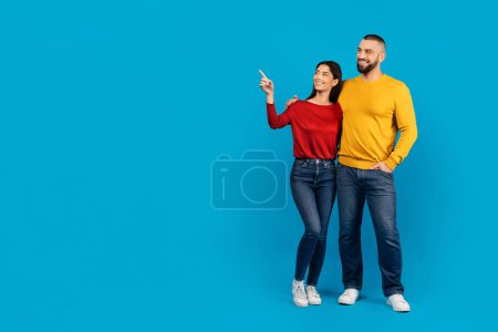 Foto de Joven pareja alegre de pie al lado del otro, mujer apuntando hacia arriba con emoción mientras el hombre mira con sonrisa, cónyuges felices posando sobre el vibrante fondo azul del estudio, longitud completa - Imagen libre de derechos