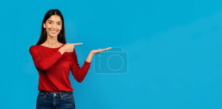 Woman wearing red shirt pointing at unknown object or direction with her hand, happy female showcasing gesture of interest or attention, standing on blue studio background, copy space