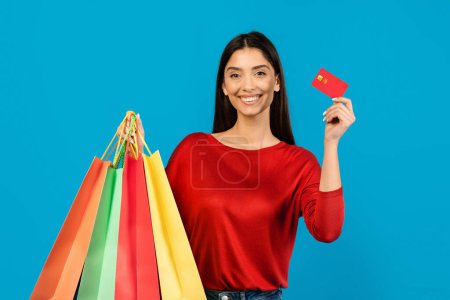 Mujer joven feliz sosteniendo bolsas de compras en una mano y tarjeta de crédito en la otra, hembra sonriente disfrutando de hacer compras, de pie sobre fondo azul estudio, espacio libre