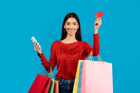 Happy woman holding multiple shopping bags, credit card and smartphone in hands, cheerful female enjoying making purchases, standing on blue background