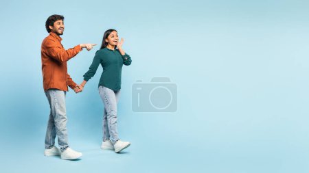 Téléchargez les photos : Plan complet de joyeux beau couple indien dans casual tenant la main et regardant de côté, pointant du doigt l'espace vide pour la publicité sur fond bleu en studio, panorama - en image libre de droit