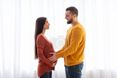 A pregnant woman and man stand closely with their hands on her belly, expressing anticipation