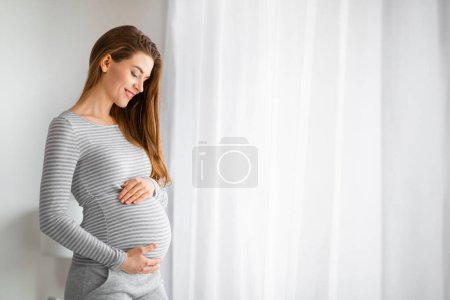 Téléchargez les photos : Femme enceinte en rayures souriant les yeux fermés, exprimant tranquillité et bonheur - en image libre de droit