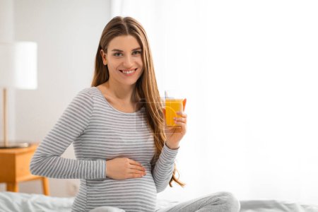 A happy pregnant woman in a striped shirt holds a glass of orange juice, posing in a well-decorated bedroom, symbolizing healthy lifestyle choices
