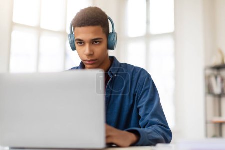Photo for Concentrated african american male teenager wearing navy blue shirt and headphones studying remotely on laptop in bright, sunlit room at home - Royalty Free Image