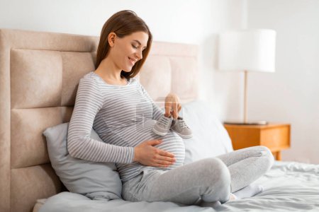 Foto de Una feliz madre embarazada con una camisa a rayas sostiene pequeños zapatos sobre su vientre, mostrando anticipación y felicidad - Imagen libre de derechos