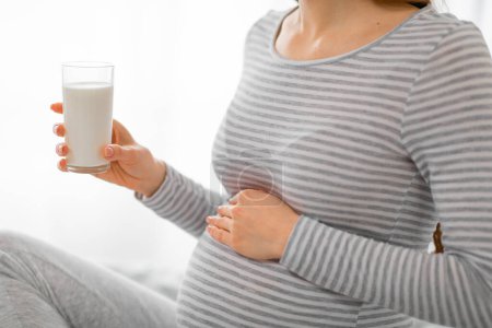 Foto de La imagen captura a una mujer embarazada vestida a rayas, sosteniendo un vaso de leche, enfatizando la importancia del calcio durante el embarazo - Imagen libre de derechos