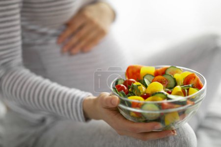 Téléchargez les photos : Gros plan d'une femme enceinte tenant un bol rempli de salade colorée, favorisant un mode de vie sain pendant la grossesse - en image libre de droit