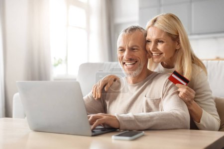 Alegre pareja madura sosteniendo una tarjeta de crédito, haciendo una compra en línea juntos en un ordenador portátil