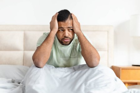 Foto de Un hombre afroamericano molesto que sufre de dolor de cabeza o estrés mientras está sentado en la cama - Imagen libre de derechos