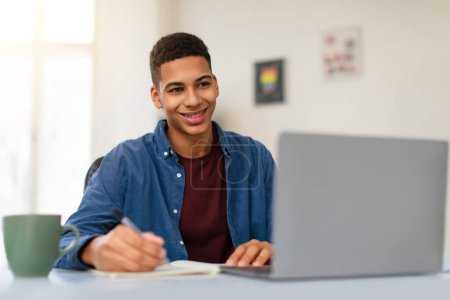 Téléchargez les photos : Heureux adolescent afro-américain gars en chemise bleue étudier avec ordinateur portable gris, prendre des notes, profiter de l'apprentissage à la maison environnement, espace libre - en image libre de droit