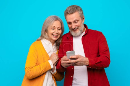 Photo for Happy mature couple casually browsing and typing messages on mobile phone, using gadget together, enjoying digital communication in their golden years, against blue background, studio shot - Royalty Free Image