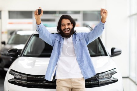 Photo for Indian man with long hair and beard joyfully raising his arms in the air in front of a new white car at a dealership - Royalty Free Image