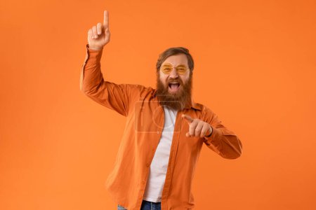 Photo for Excited redhaired bearded man wearing sunglasses, dances with cool vibes having fun against orange studio backdrop. Concept of partying and joyful energy of movement and celebration - Royalty Free Image