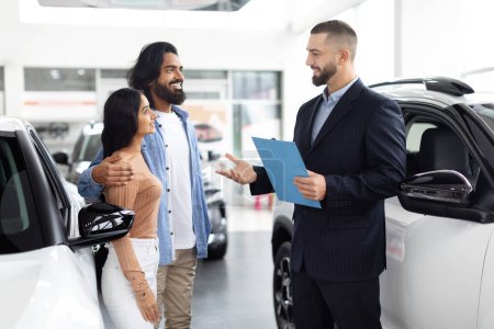 Smiling car salesman has a friendly chat with eastern couple by a white car in a showroom