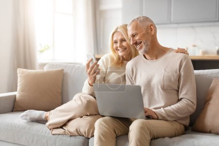 Téléchargez les photos : Couple âgé assis sur le canapé, partageant un rire en regardant un écran d'ordinateur portable, dans un salon bien éclairé - en image libre de droit