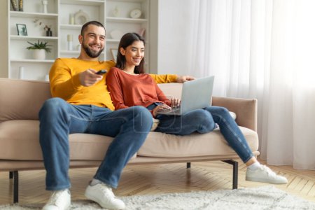 Téléchargez les photos : Un couple joyeux profite du temps libre sur un canapé, l'un avec un ordinateur portable et l'autre tenant une télécommande - en image libre de droit