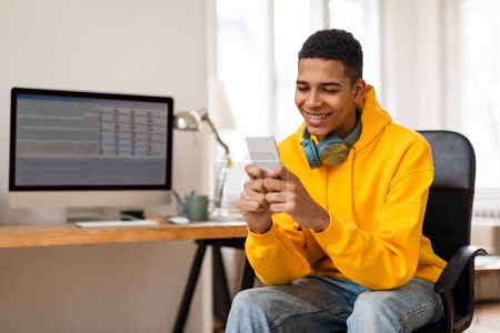 Téléchargez les photos : Un jeune homme joyeux portant un sweat à capuche jaune utilise son smartphone avec un écran d'ordinateur en arrière-plan - en image libre de droit