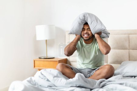 An irritated african american man tries to block out noise by covering his ears with a pillow, indicating a need for quiet