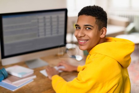 Téléchargez les photos : Heureux adolescent noir en sweat à capuche jaune se tournant pour sourire à la caméra tout en étudiant sur ordinateur à la maison, montrant de la confiance et de la joie, profiter de e-learning - en image libre de droit