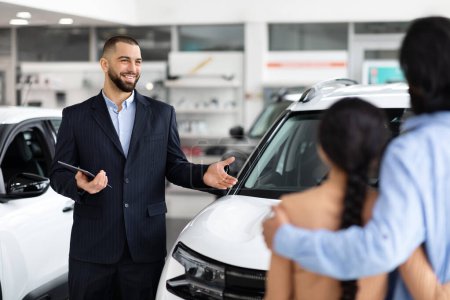 A car salesman points out features of a white vehicle to a hindu couple in a dealership, with focus and attention