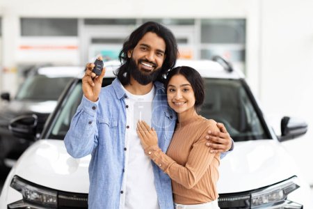Ein glückliches junges indisches Paar mit Autoschlüssel vor einem Neuwagen in einem Autohaus, symbolisiert einen Neukauf